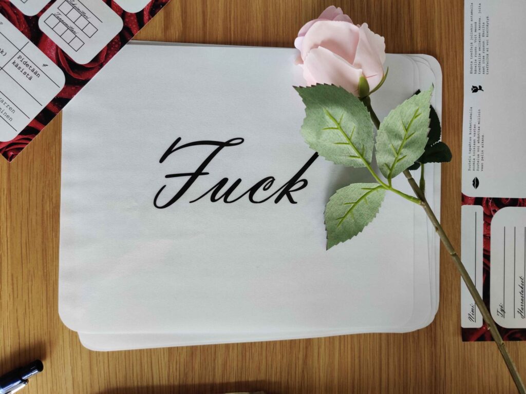 Photo of a tabletop with a plastic pink rose laying on top of a white sign with "Fuck" on it in cursive. There are partially visible character sheets beside the sign.