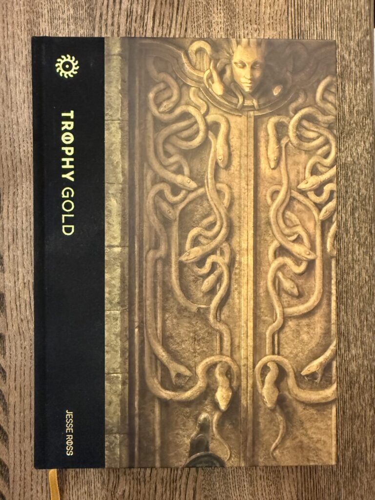 Cover of Trophy Gold. It's gold and black, depicting massive double doors with a Medusa head on top in relief. A lone figure with a helmet and a shield is facing the doors, almost out of picture.
