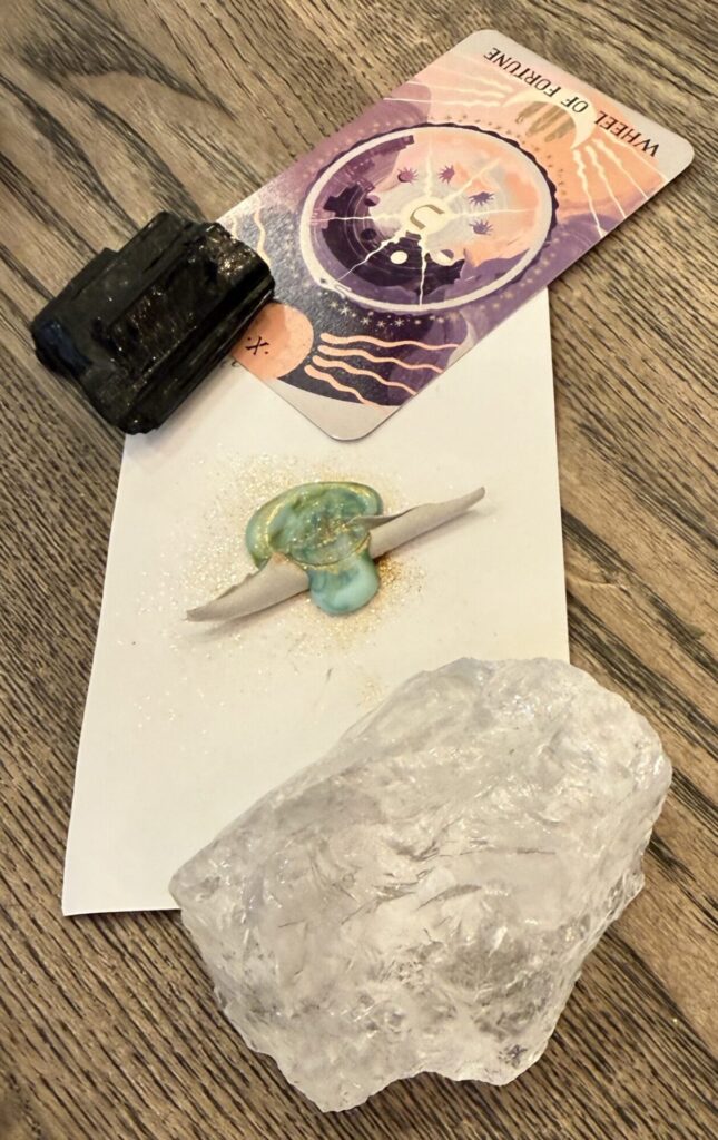 A folded piece of paper with a wax seal. There's glitter on the wax. Rare stones are on top, a translucent one and a shiny black one. A tarot card, The Wheel of Fortune, is laid on top.